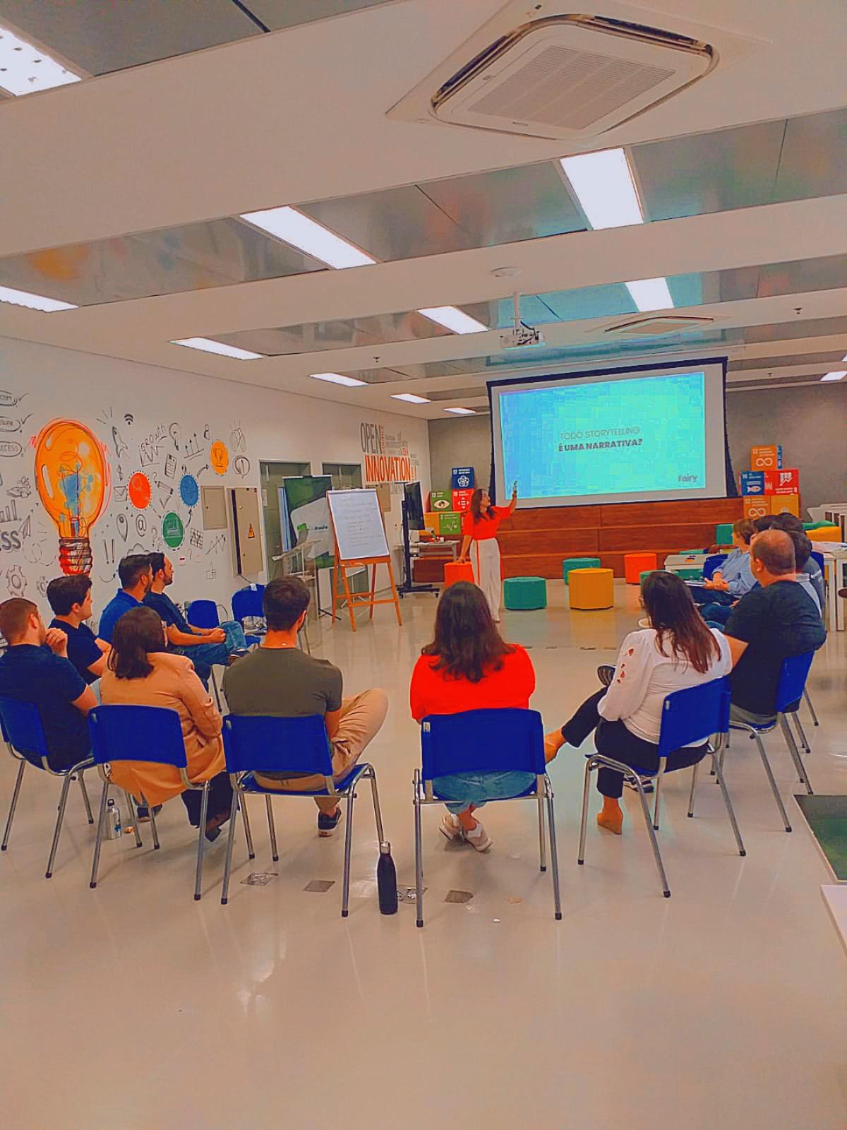 Taís Delduck palestrando para um grupo sentado em roda, com a apresentação projetada ao fundo